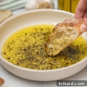 Toasted baguette being dipped into olive oil bread dip with fresh garlic in background.