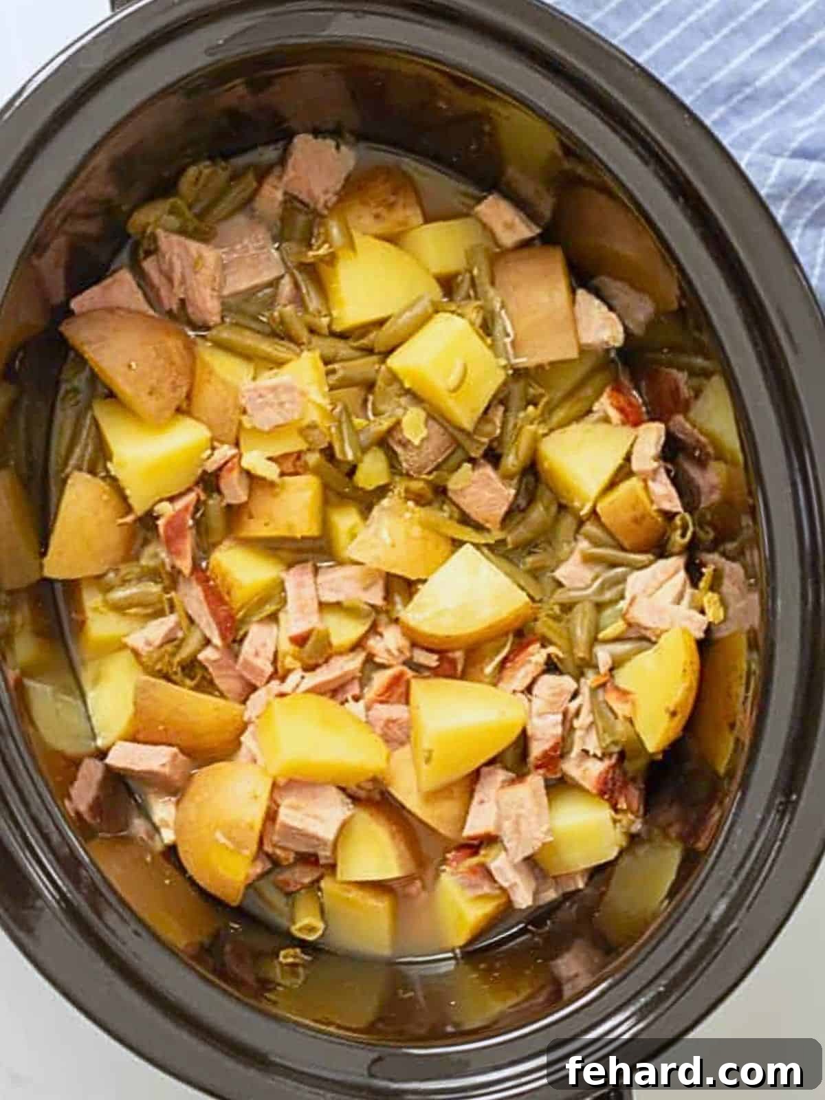 A close-up shot of tender Yukon gold potatoes, vibrant green beans, and savory ham chunks, all perfectly cooked together in a slow cooker, ready to serve as a comforting meal.