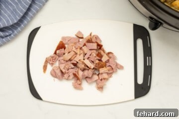 Diced ham on a clean white cutting board, positioned near the slow cooker in the background, ready to be stirred back into the dish.