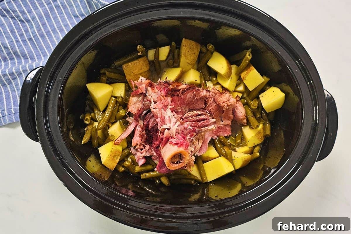 Inside a slow cooker, a ham bone is nestled among chunky potatoes and vibrant green beans, all seasoned and ready for the slow cooking process.