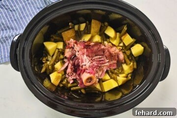 Slow cooker with ham bone, potatoes, and green beans inside, seasoned and ready to cook.