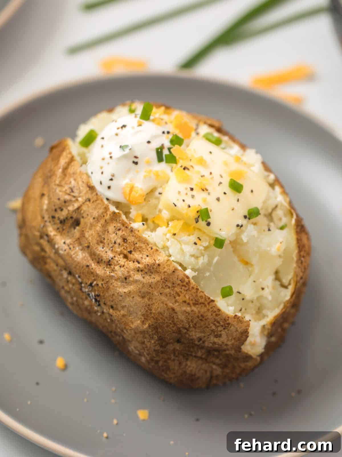 Air Fryer Baked Potato Recipe A perfectly air fried baked potato on a gray plate, cut open and lavishly topped with a dollop of butter, creamy sour cream, shredded cheddar cheese, a sprinkle of black pepper, and fresh chives.