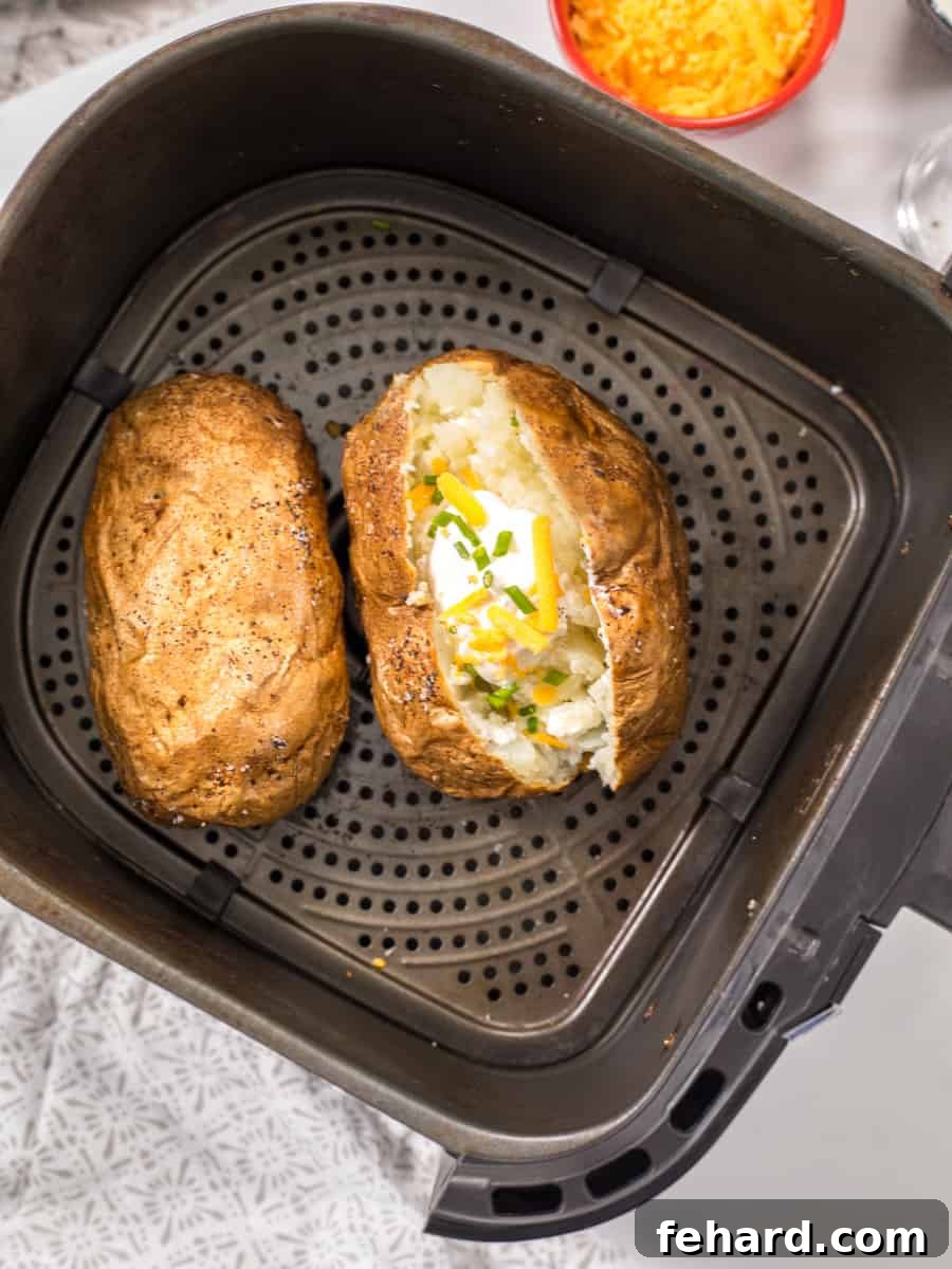 Baked Potatoes Air Fryer Two large, inviting baked potatoes, fresh from the air fryer, presented on a gray plate. One potato is expertly cut open and generously adorned with a dollop of butter, a scoop of sour cream, melted cheddar cheese, a dash of freshly ground black pepper, and vibrant green chives, ready for enjoyment.
