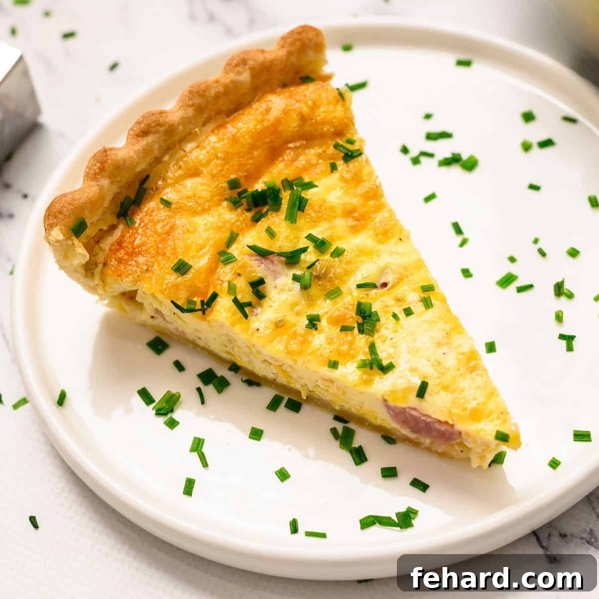 Ham & Cheese Quiche Recipe A perfect slice of ham and cheese quiche, garnished with fresh chives, presented on a pristine white plate.
