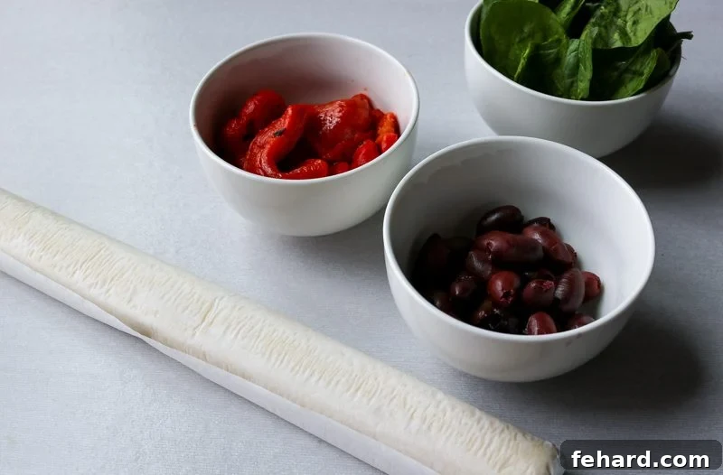 Ingredients for Greek Rolls Fresh ingredients laid out for Greek Pinwheels, including puff pastry, spinach, olives, roasted red peppers, and feta cheese.