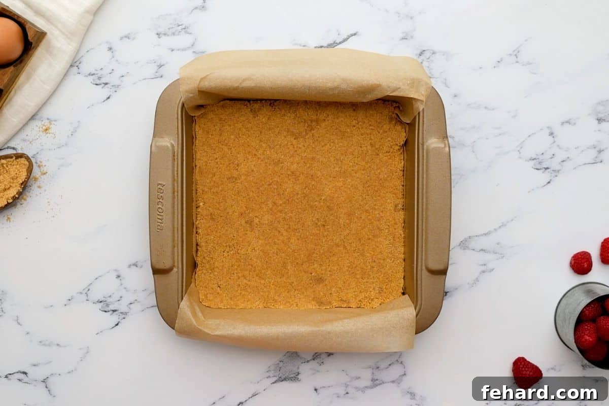 Graham Cracker crust pressed into an 8x8 baking dish lined with parchment paper with raspberries in background.