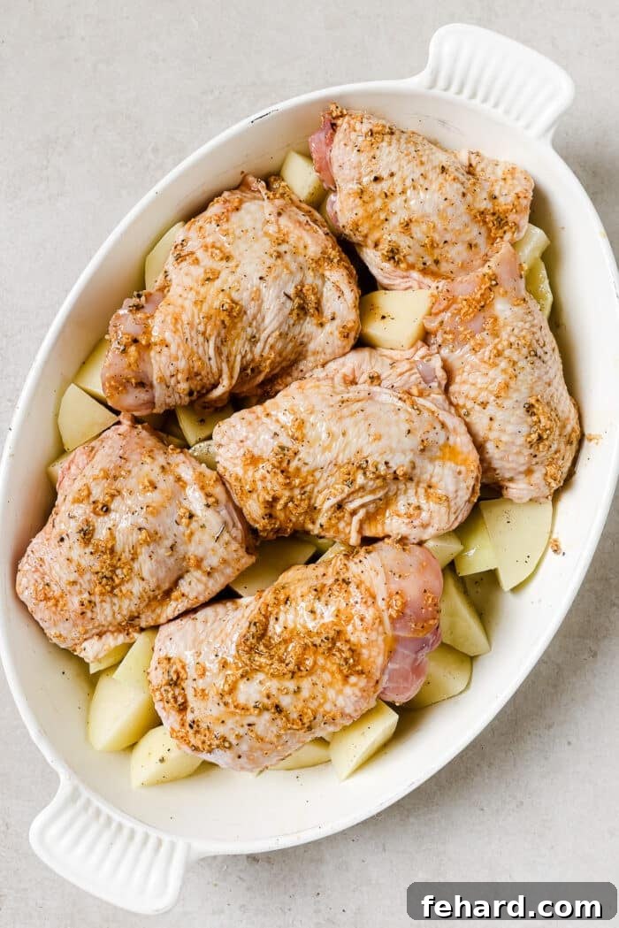An overhead view of marinated chicken thighs over diced potatoes in a white baking dish.