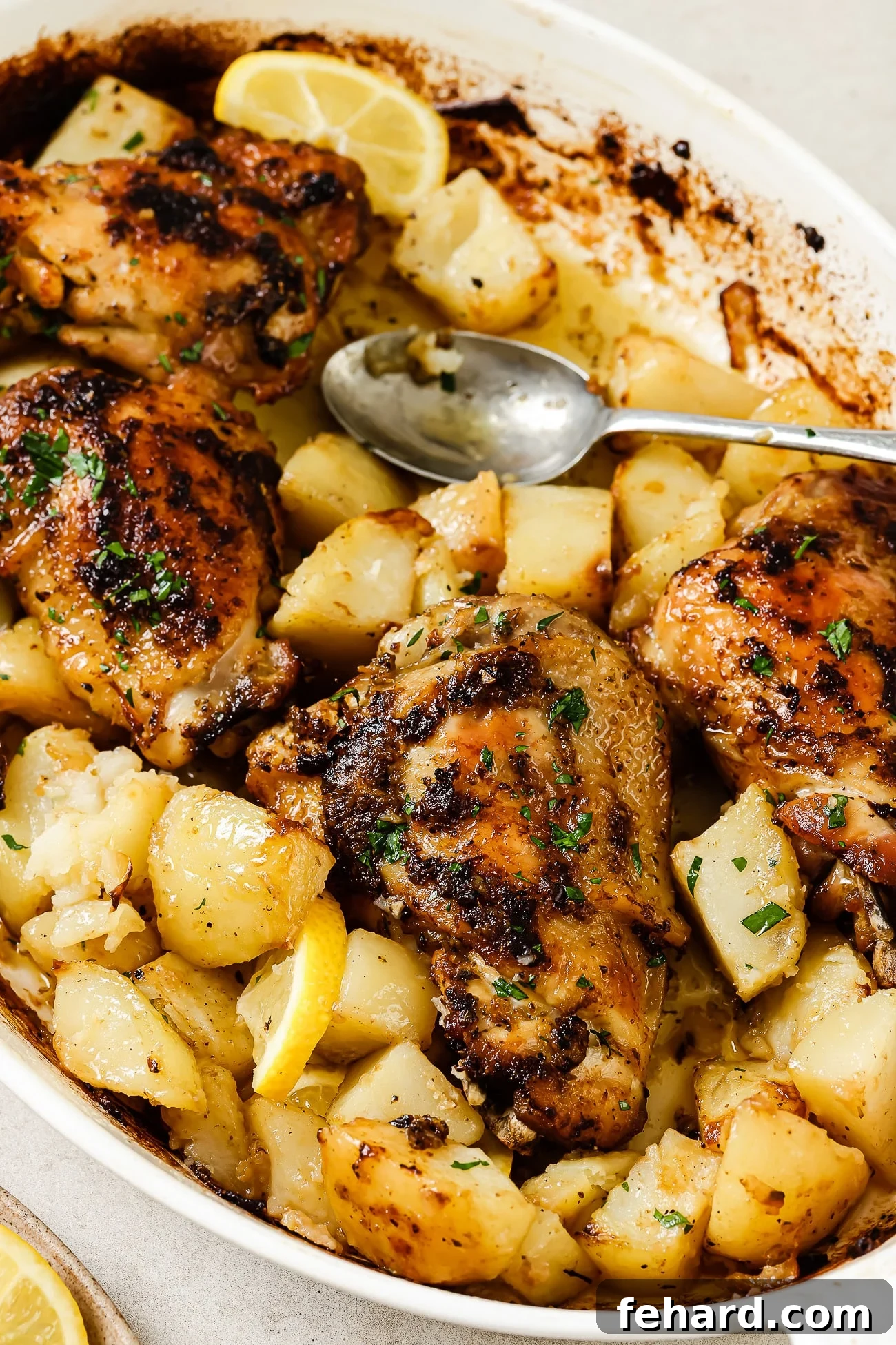 Baked Greek chicken and potatoes in a white baking dish with a serving spoon in it.