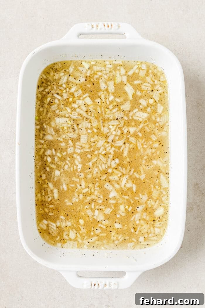 A white casserole dish filled with uncooked long-grain rice, diced onions, minced garlic, olive oil, melted butter, and hot liquids before adding chicken.