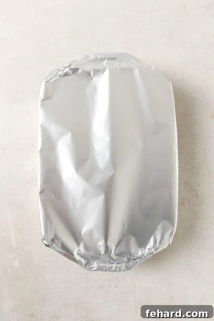 An overhead view of a white casserole dish, tightly covered with aluminum foil, ready for the oven.
