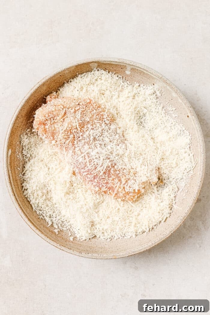A chicken cutlet in a bowl of breadcrumbs and parmesan cheese, being pressed to ensure full coating.