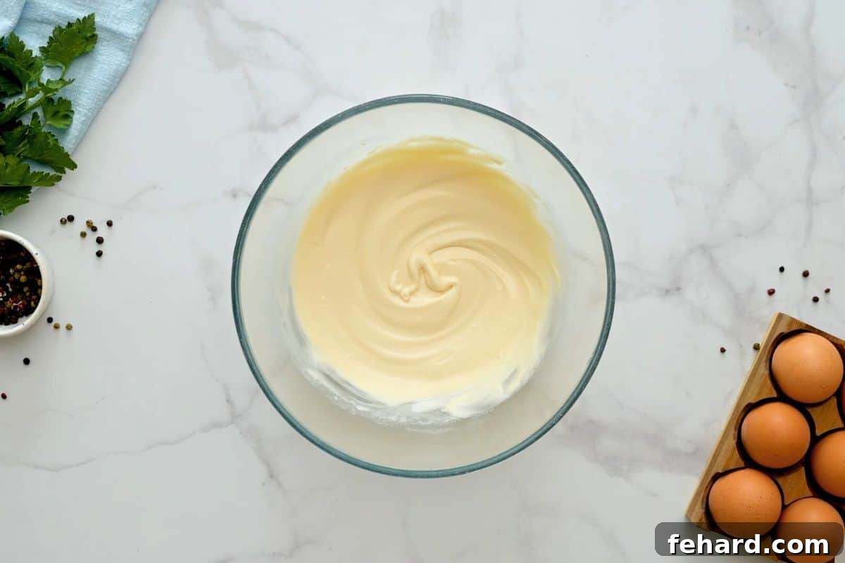 Hellmann's mayonnaise being whisked with white vinegar and kosher salt in a large glass mixing bowl, forming the creamy potato salad dressing.