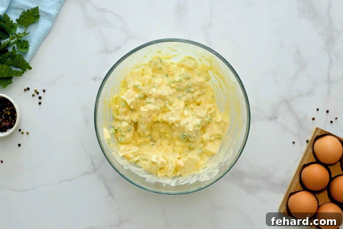 A large mixing bowl containing cooked potatoes, hard-boiled eggs, celery, and onions, all gently mixed into a creamy mayonnaise-based dressing.