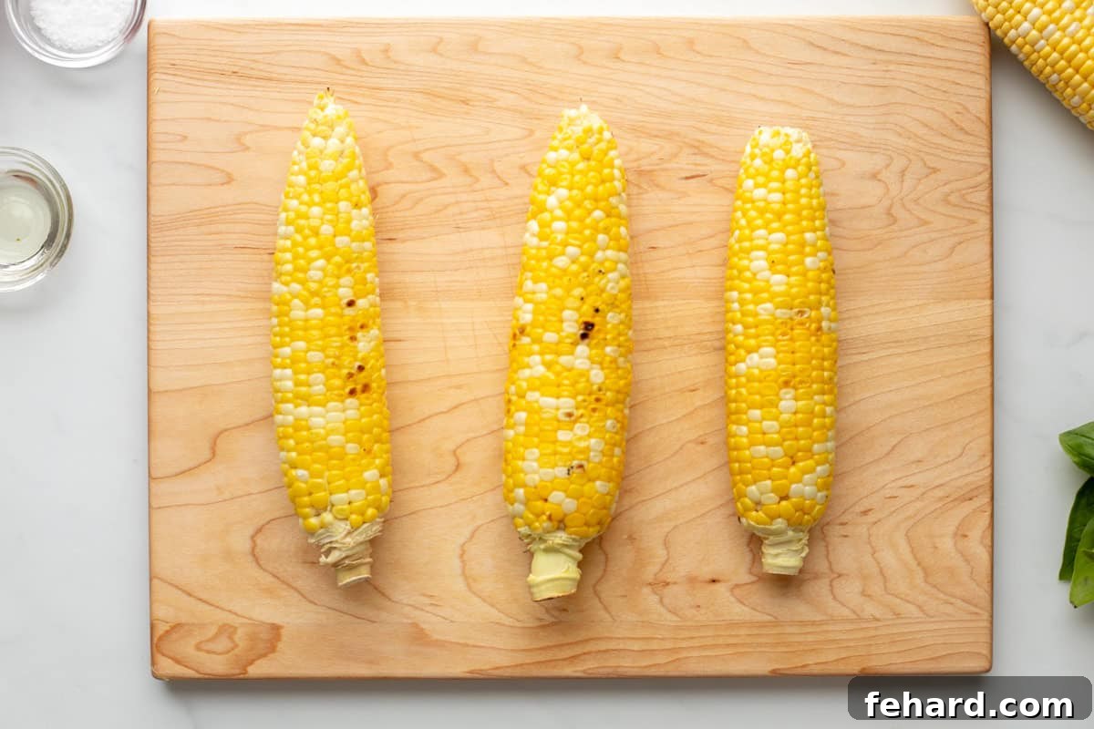 Three perfectly grilled ears of corn, showing golden kernels with slight char marks, resting on a rustic wooden cutting board, ready for the next step in making corn and tomato salad.