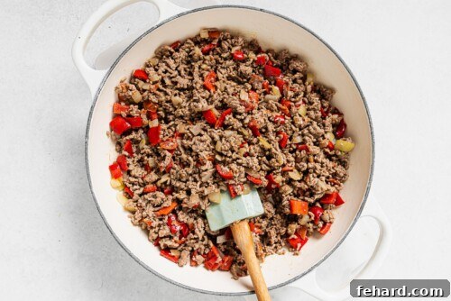 Browned ground beef cooking with sautéed onions, bell pepper, and tomato in a white skillet, ready for the sauce.
