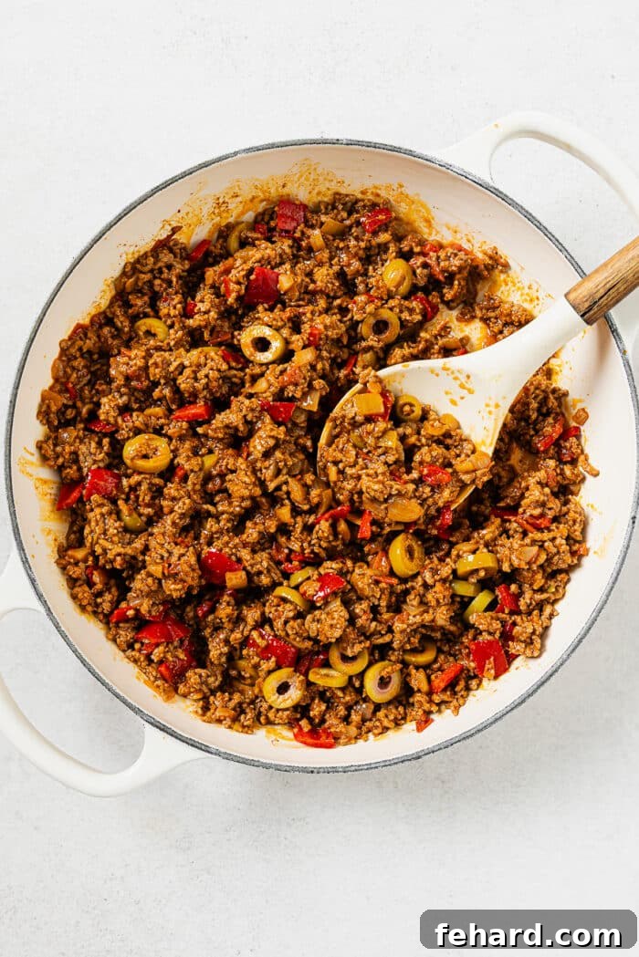 A large pot of simmering Picadillo, rich with ground beef, vegetables, and sauce, featuring a white serving spoon ready for dishing.