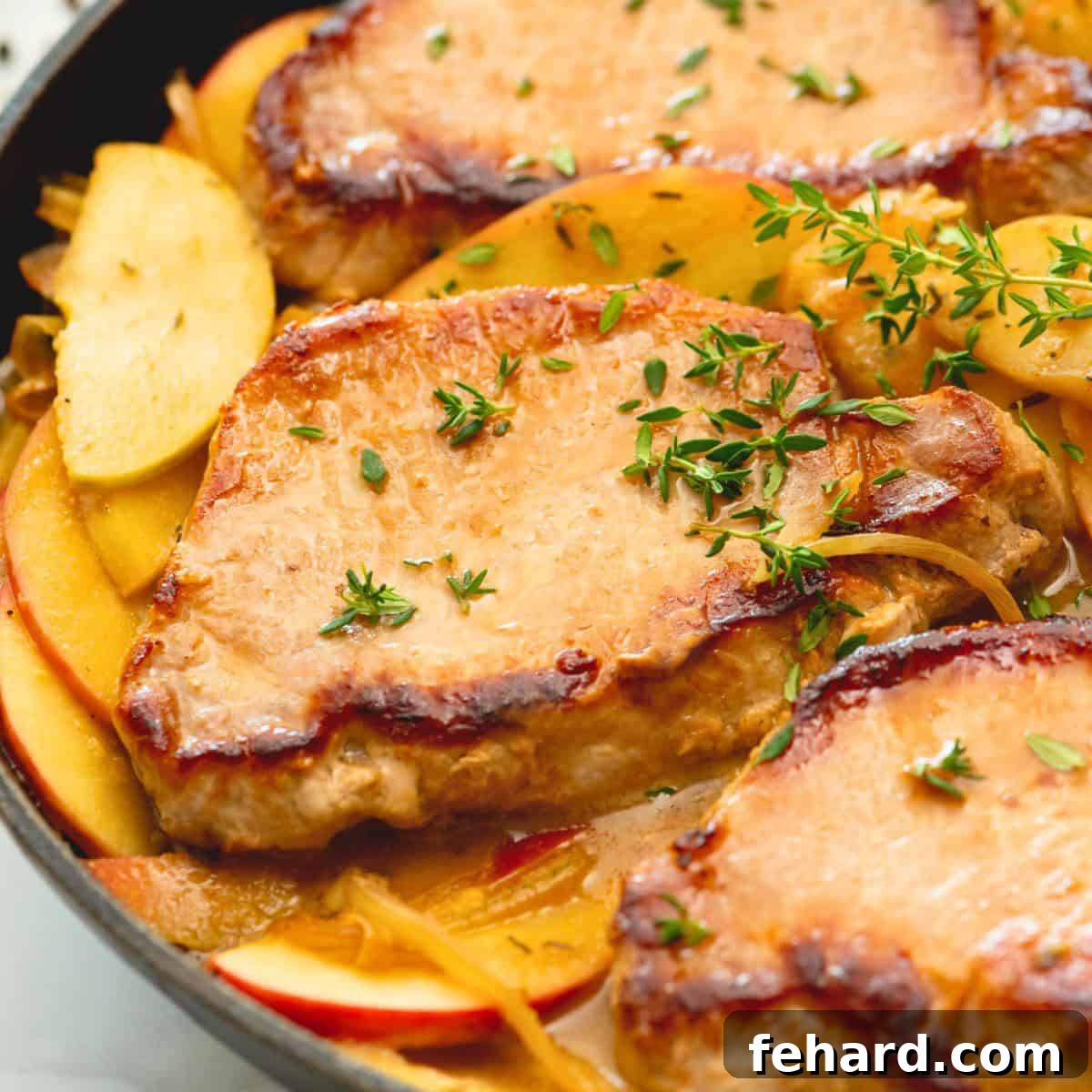 Pork Chops with Apples and Onions Skillet Close up of perfectly seared pork chops with glistening caramelized apples and onions in a sauté pan, garnished with fresh thyme leaves.