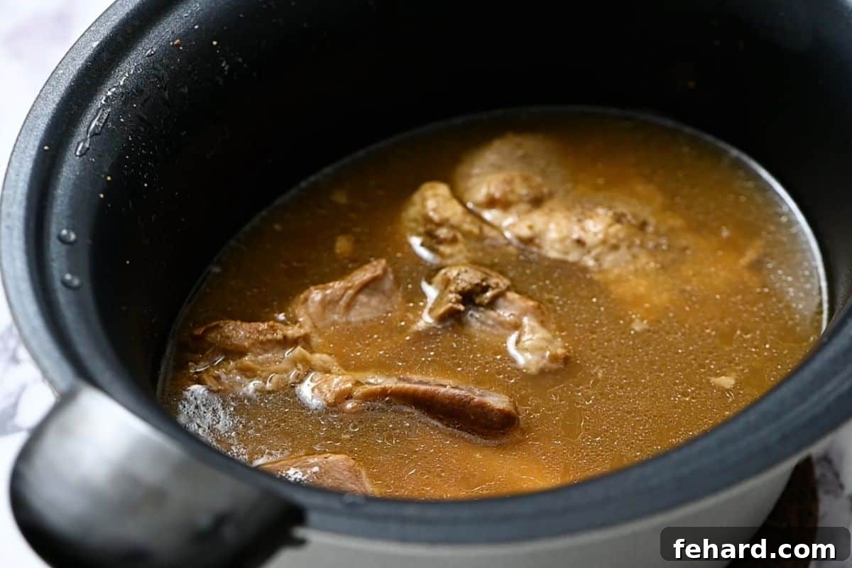 Cooked pork shoulder chunks swimming in a rich, flavorful braising liquid inside a slow cooker, appearing perfectly tender after hours of slow cooking.