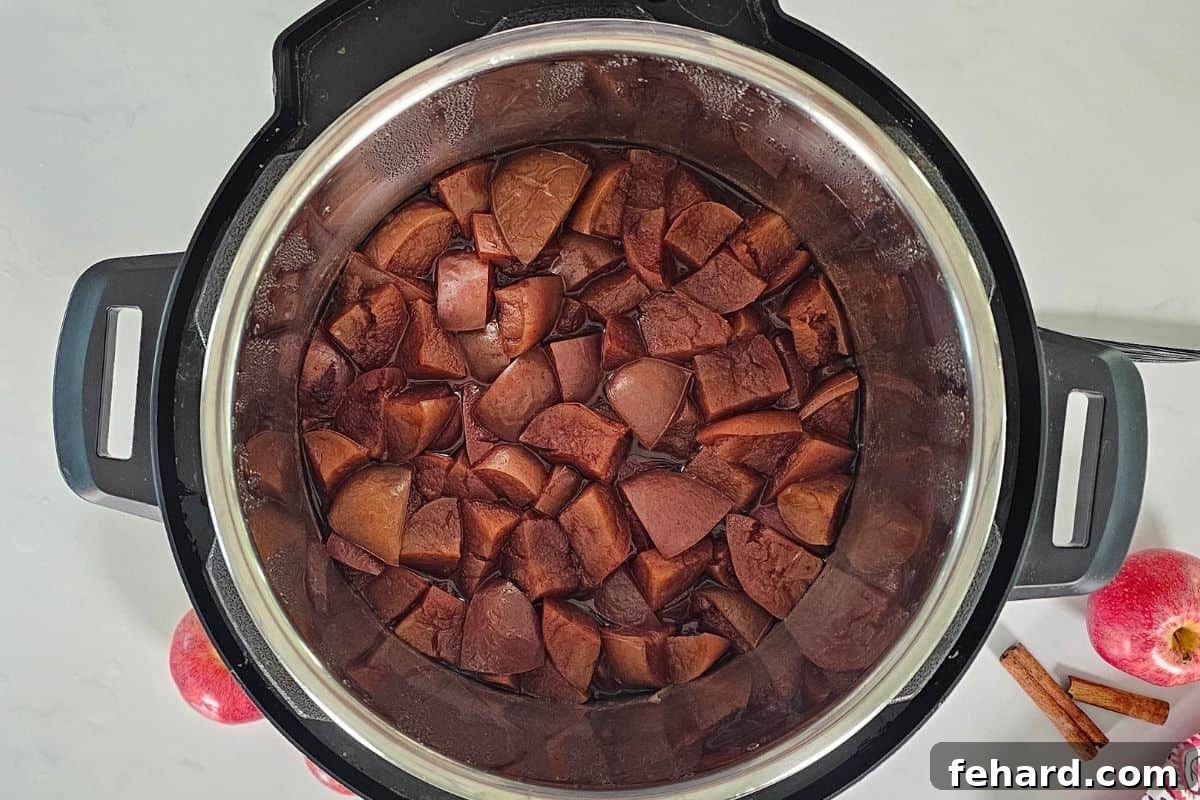 IP Apple Butter Cooked apples in the inner pot of a pressure cooker next to fresh apples and cinnamon sticks, showcasing the softened fruit.