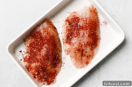 Seasoned raw tilapia fillet on a white baking sheet.