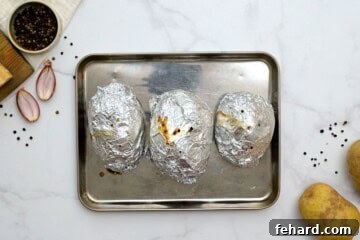 Three large russet potatoes wrapped in foil on baking sheet after being baked for potatoes romanoff casserole.