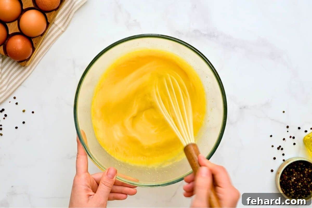 Whisking Egg Mixture A whisk vigorously mixing 12 large eggs and milk in a clear glass mixing bowl, creating a smooth, yellow egg casserole base.