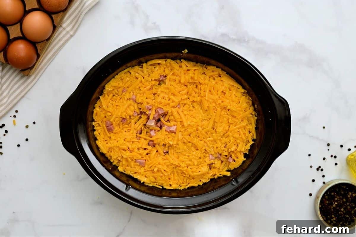 Layering Breakfast Casserole in Crockpot Close-up of a large slow cooker filled with distinct layers of frozen hash browns, diced ham, vibrant peppers and onions, and shredded cheese, with the whisked egg mixture being poured over the top.