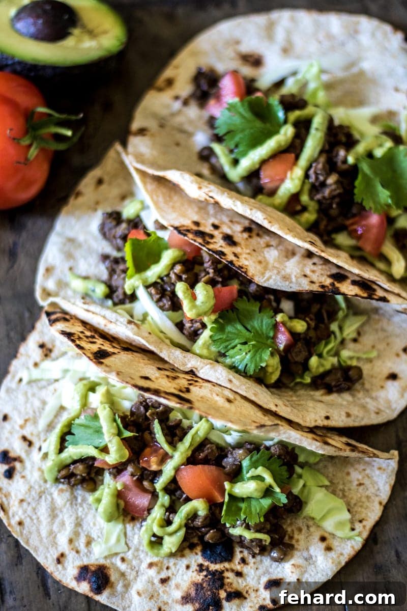 Three perfectly arranged lentil tacos, generously topped with creamy avocado-lime sauce and fresh shredded cabbage, presented on a wooden cutting board.