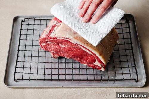 Patting dry a raw prime rib roast with a paper towel on a wire rack to remove moisture before roasting for the reverse sear prime rib.
