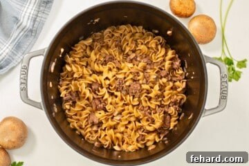 Perfectly cooked egg noodles and ground beef in large Dutch Oven next to fresh mushrooms.