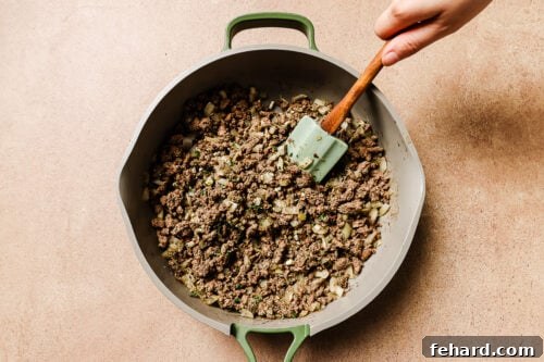 A hand stirs cooked ground beef and chopped onions in a large, light gray skillet with green handles using a green spatula, preparing the savory base for Baked Rigatoni with Ground Beef on a light brown surface.
