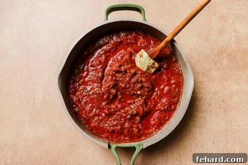 A green-handled pot filled with chunky red tomato sauce for Baked Rigatoni with Ground Beef sits on a beige surface. A wooden spatula rests in the thick, hearty sauce, rich with visible tomato and herb pieces.