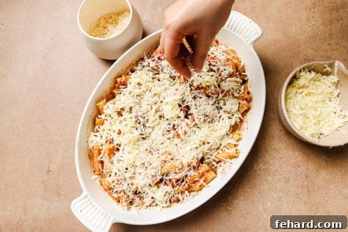 A hand sprinkling mozzarella cheese on top of the rigatoni with ground beef in a casserole dish.
