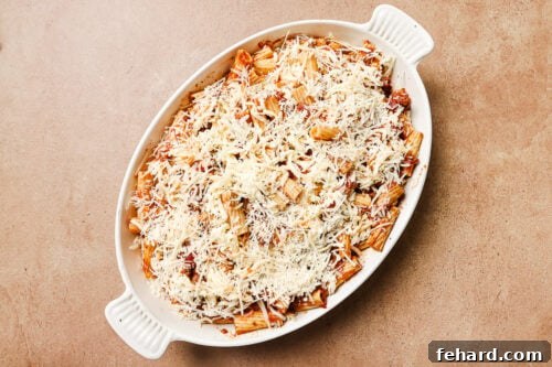 A white oval baking dish filled with Baked Rigatoni with Ground Beef, mixed with tomato sauce and topped with a generous layer of shredded cheese, ready to be baked. The dish sits on a light brown surface.