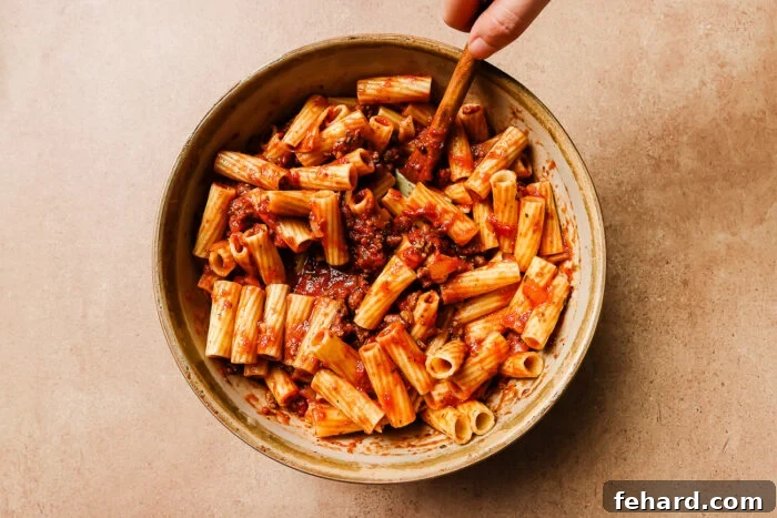 A large bowl where cooked rigatoni pasta is being expertly tossed with the rich ground meat sauce, ensuring every tube is thoroughly coated.