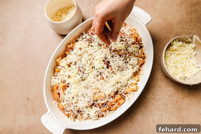 A hand generously sprinkling shredded mozzarella cheese over the first layer of rigatoni with ground beef in a casserole dish.