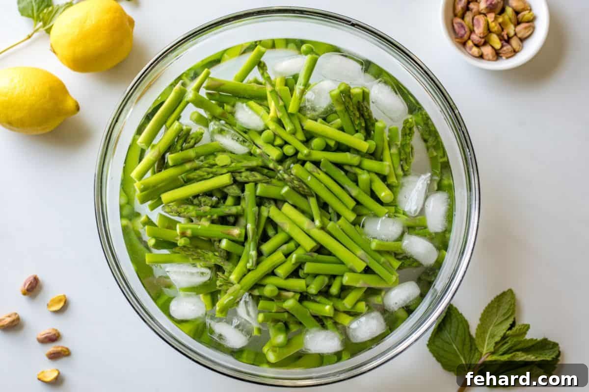 Blanched asparagus and sweet peas cooling in a bowl of ice water, with fresh mint leaves and chopped pistachios in the background, ready to be added to the salad.