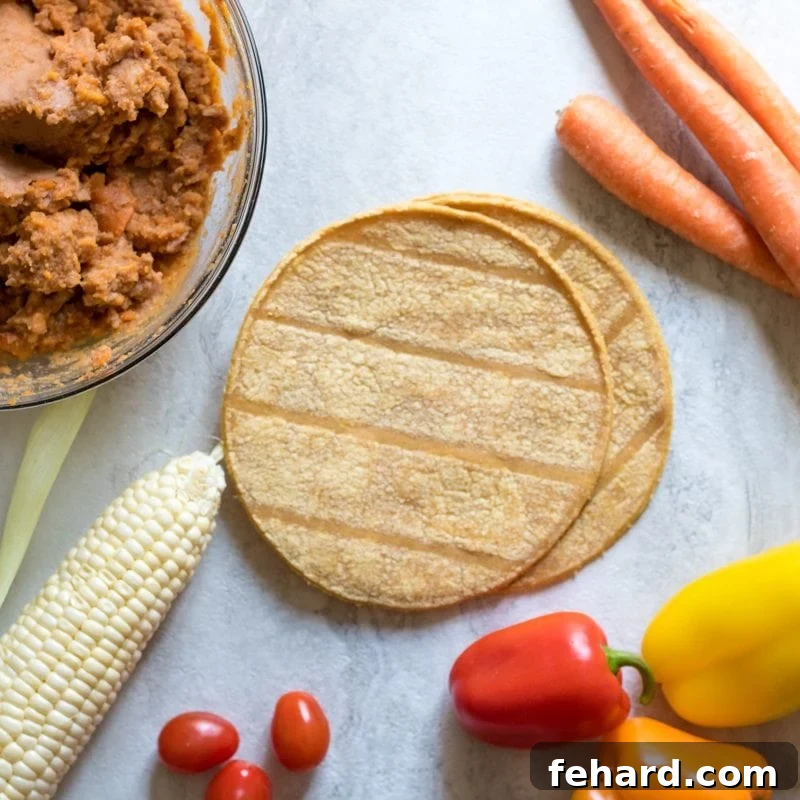 Baked Tostada Ingredients Fresh ingredients for homemade tostadas laid out on a white cutting board, including corn tortillas, a bowl of refried beans, shredded cheese, and various vegetables like tomatoes and peppers.
