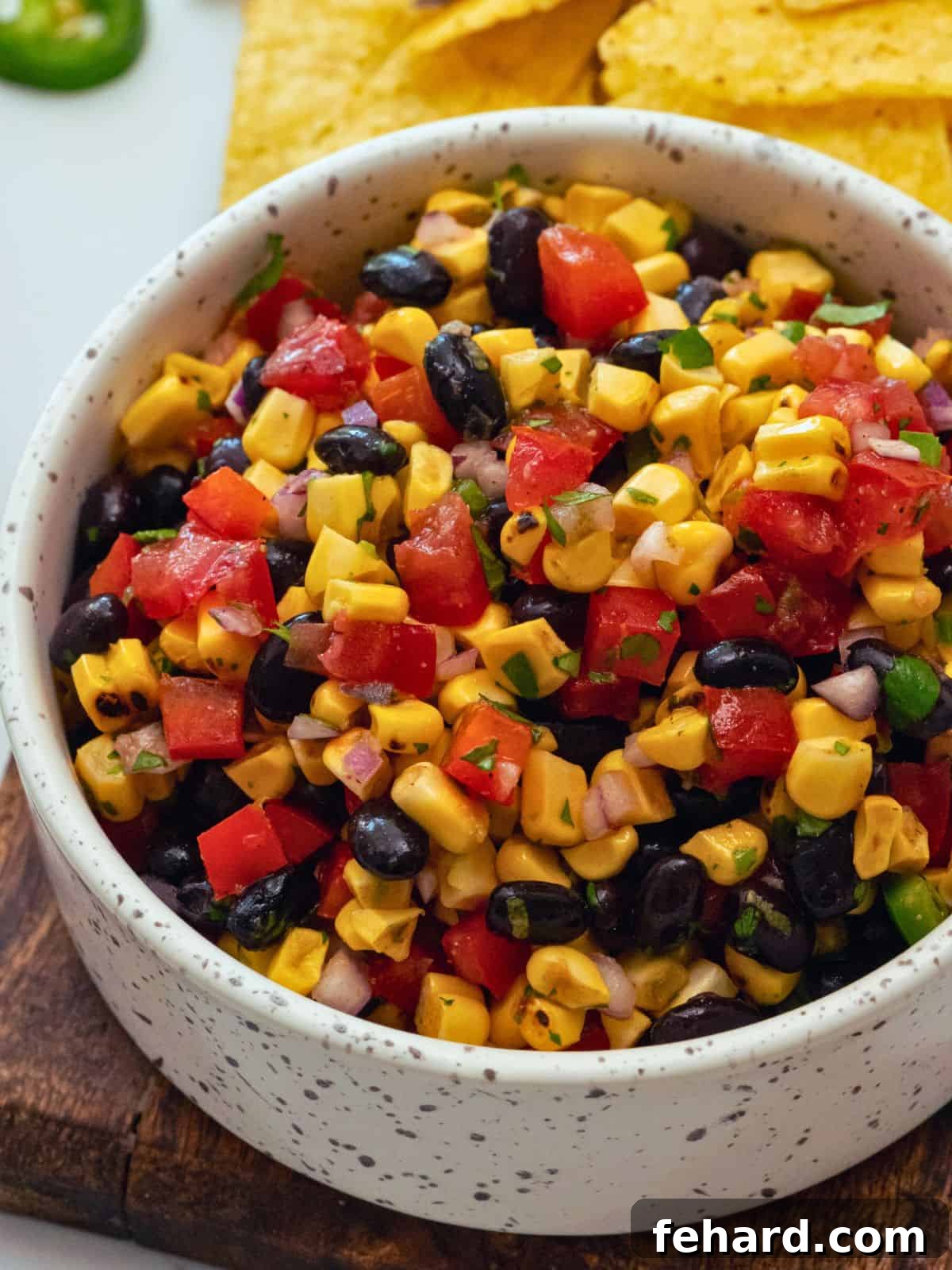 A vibrant bowl of black bean and corn salsa, featuring charred corn kernels, diced tomatoes, bell peppers, jalapeños, and fresh cilantro, served alongside golden tortilla chips.