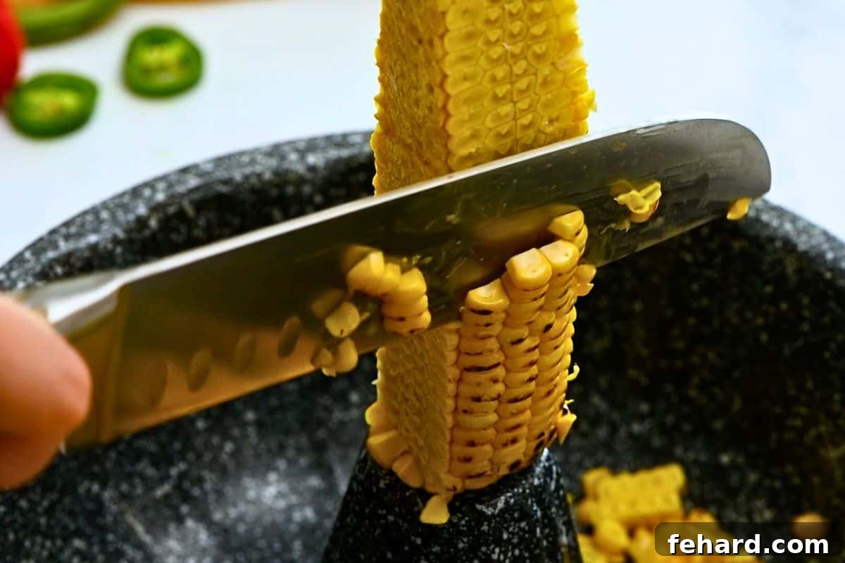 A corn cob positioned in the center of a bundt pan, with a sharp knife carefully slicing off kernels directly into the pan.