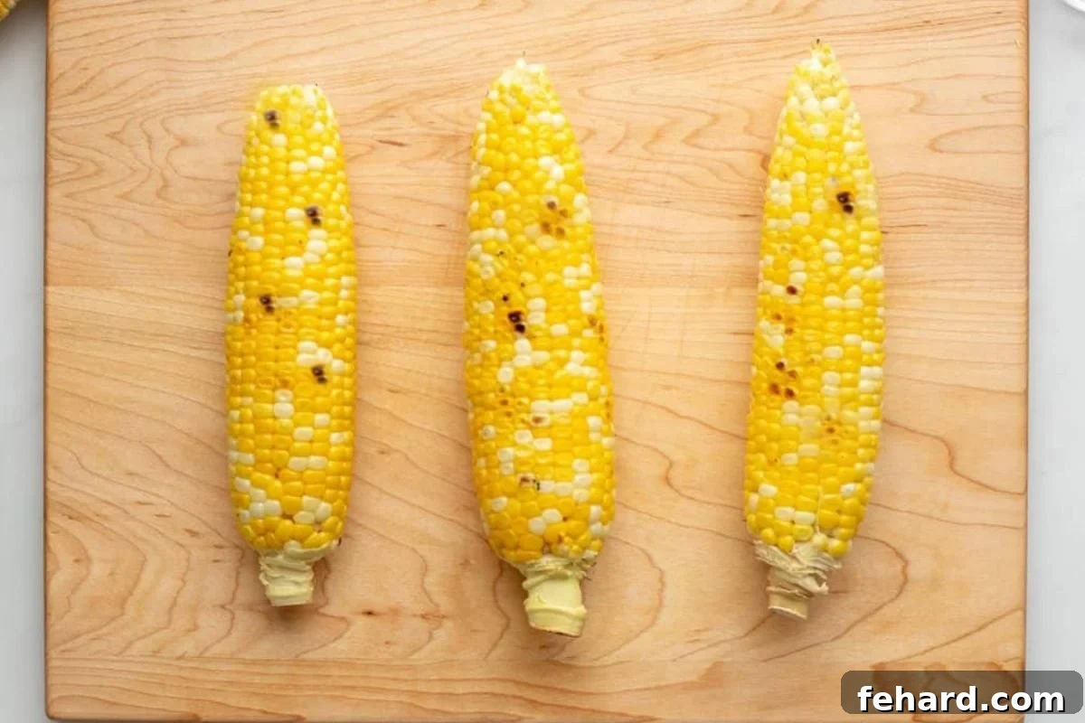 Three ears of lightly grilled corn on the cob resting on a wooden cutting board, ready for preparation.