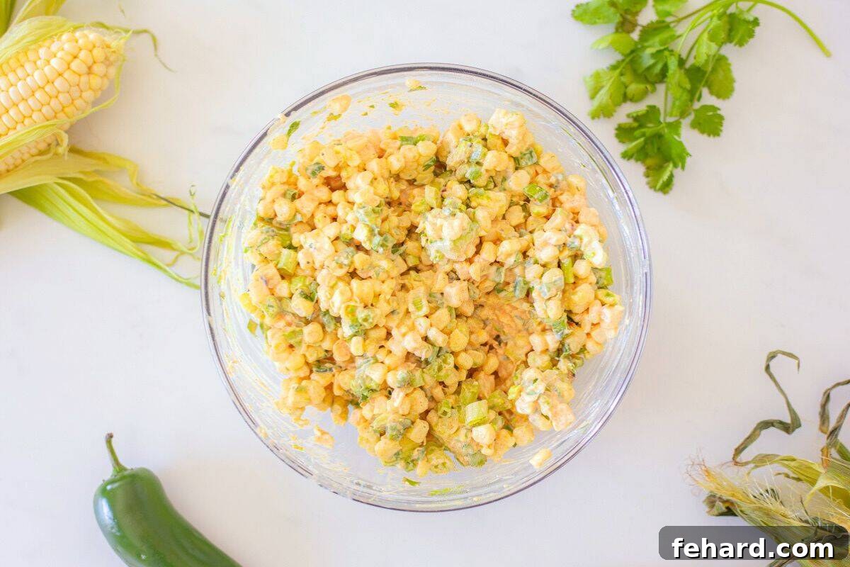 Grilled corn, diced jalapeno, sliced green onions, and cubed avocado being stirred into a creamy Greek yogurt and sour cream dressing for Mexican Street Corn Salad.