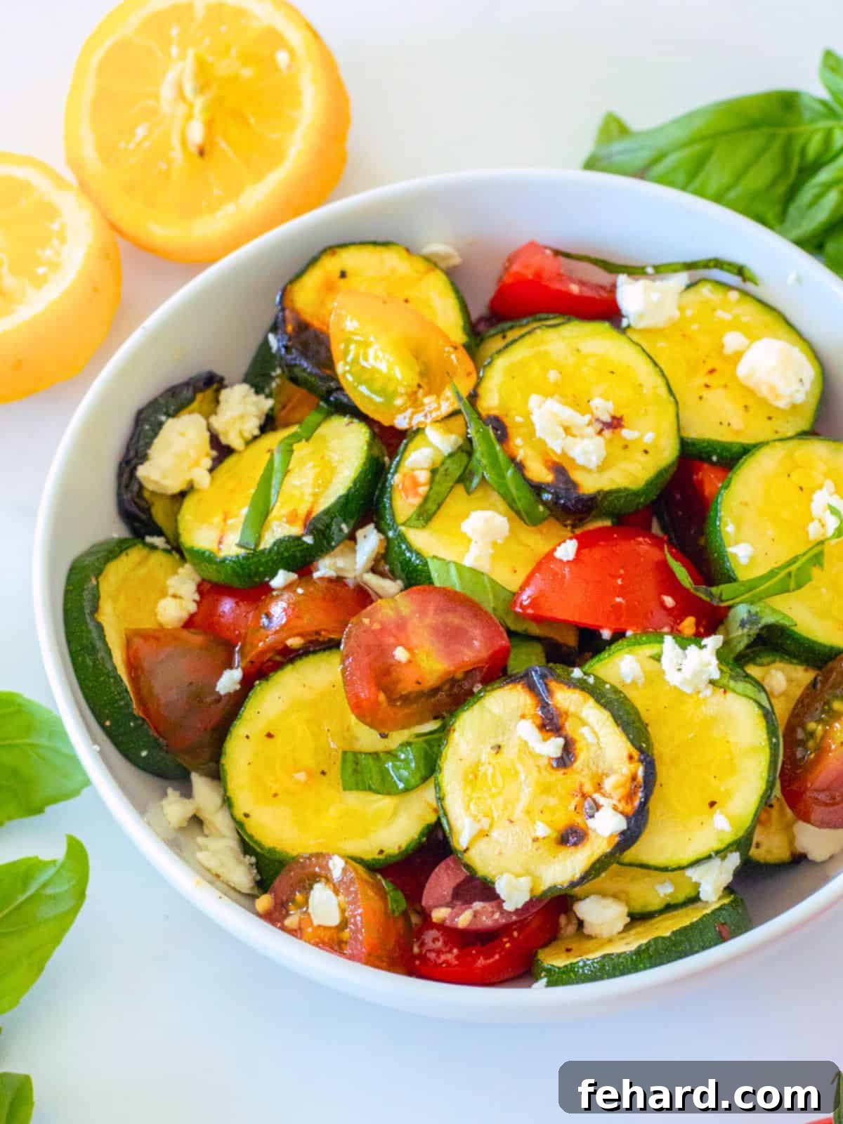 Grilled Zucchini Salad Recipe: A Summer Essential Vibrant zucchini salad with tender grilled zucchini, colorful tomatoes, and crumbled feta cheese, garnished with fresh basil in a white serving bowl. Additional fresh tomatoes and basil are blurred in the background, suggesting a garden-fresh meal.