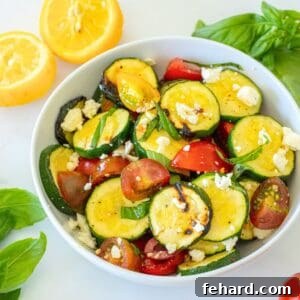 Grilled zucchini salad with fresh tomatoes and crumbled feta in a white serving bowl, garnished with basil, with a blurred background of fresh produce.