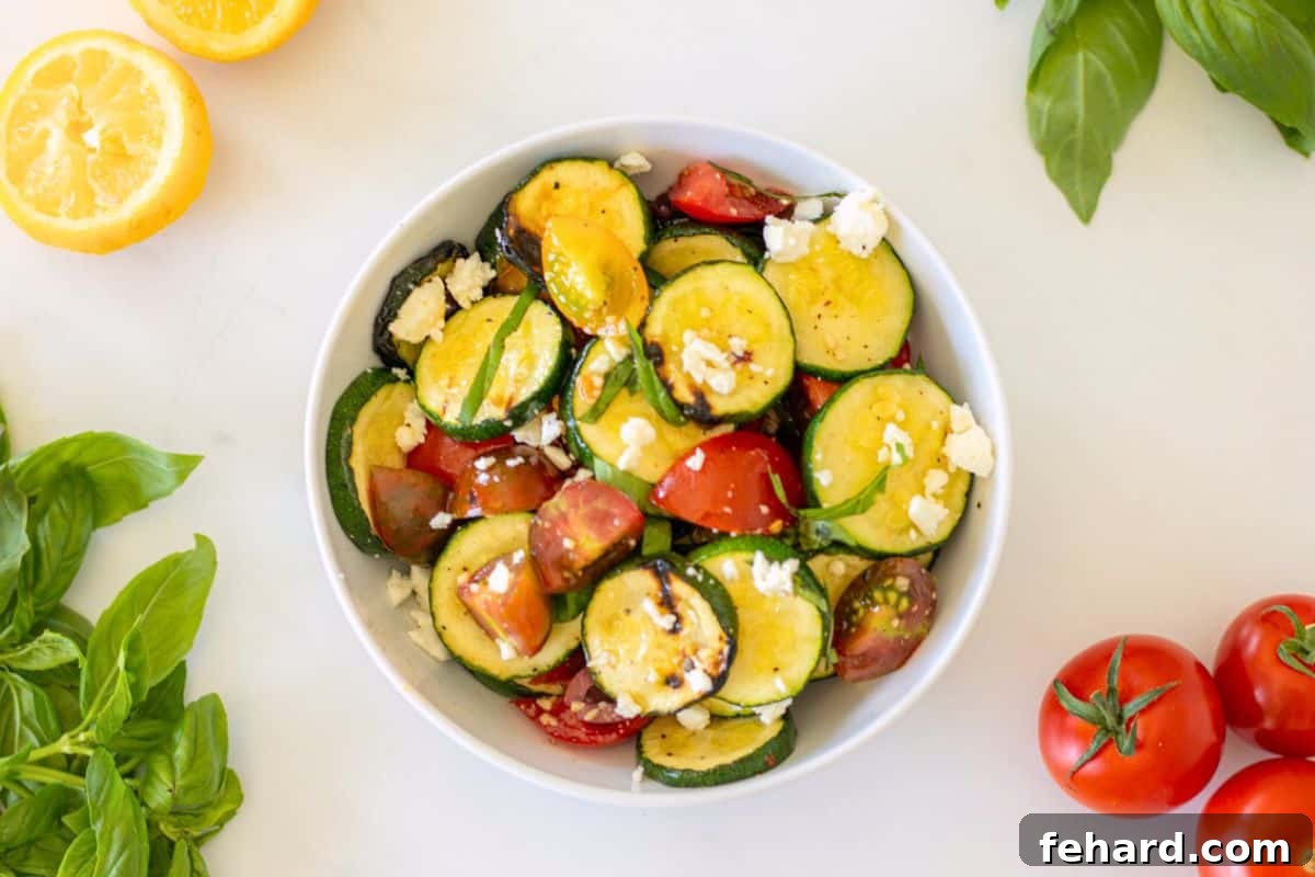 Finished Grilled Zucchini Salad Zucchini salad, beautifully presented in a white serving dish, featuring tender grilled zucchini, colorful tomatoes, and crumbled feta, garnished with fresh basil. Additional fresh tomatoes are softly blurred in the background.