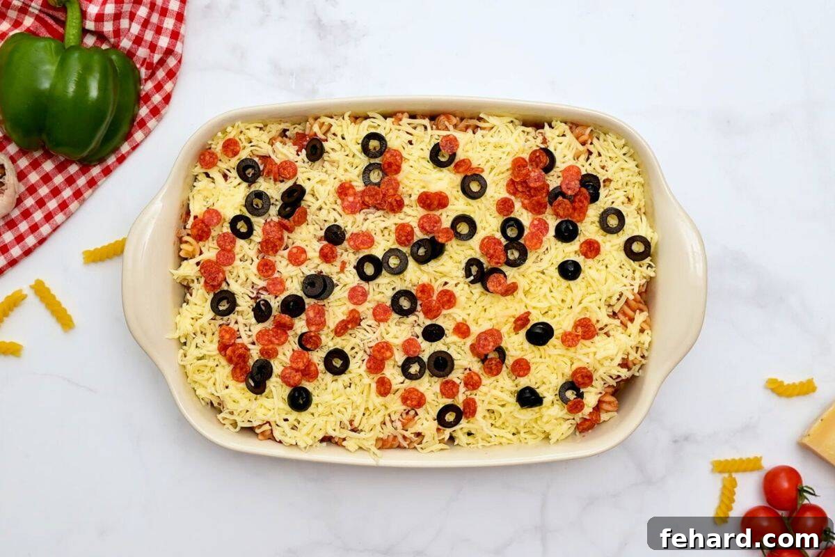 Cooked pasta layered in casserole dish topped with shredded cheese, olives, and pepperoni before baking.