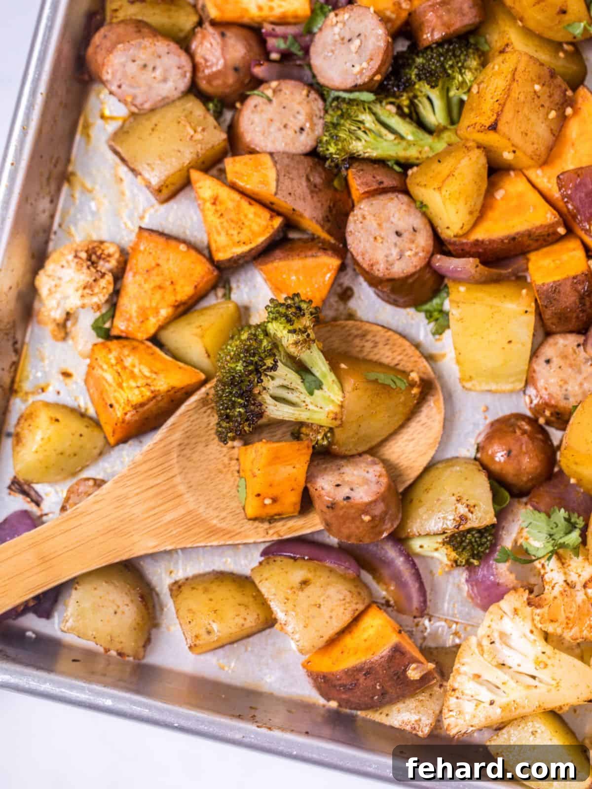 Wooden spoon scooping up perfectly roasted sweet potato, potato, broccoli, and chicken sausage off sheet pan, showcasing a delicious and easy weeknight dinner.