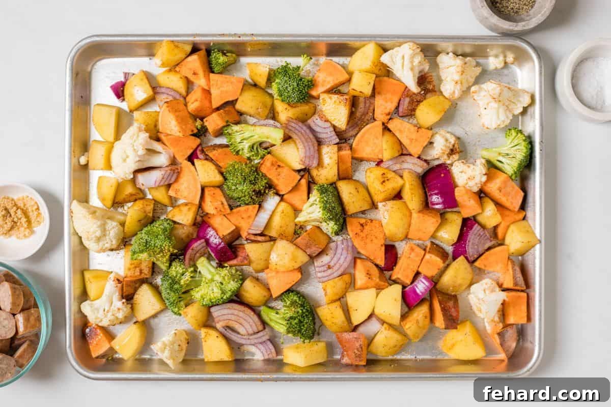Potatoes, sweet potatoes, broccoli, cauliflower, and red onions on rimmed sheet pan tossed with olive oil and seasonings, ready for roasting.