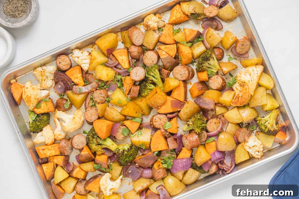 Chicken sausage with potatoes, sweet potatoes, and mixed veggies perfectly roasted on a sheet pan, with pot holders on the side, indicating it's fresh from the oven.