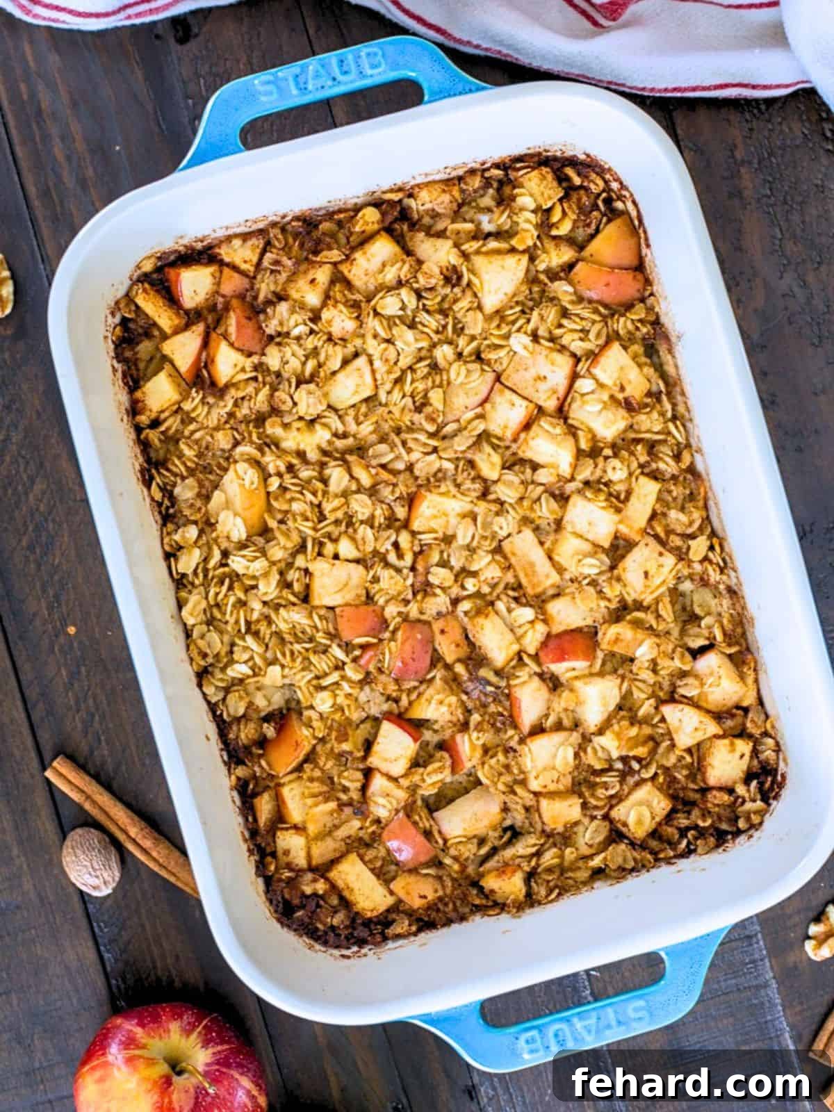 Baked apple oatmeal in a blue baking dish, garnished with walnuts and a sprinkle of cinnamon, beside cinnamon sticks and whole fresh apples.